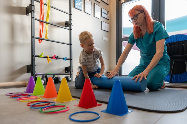 Pediatric Physiotherapist Working With Young Child Patient During Physical Rehabilitation Session In Medical Clinic. Therapist Preparing Balance And Motor Coordination Exercise Using Foam Roller, Cones And Rings For Child Development Therapy.